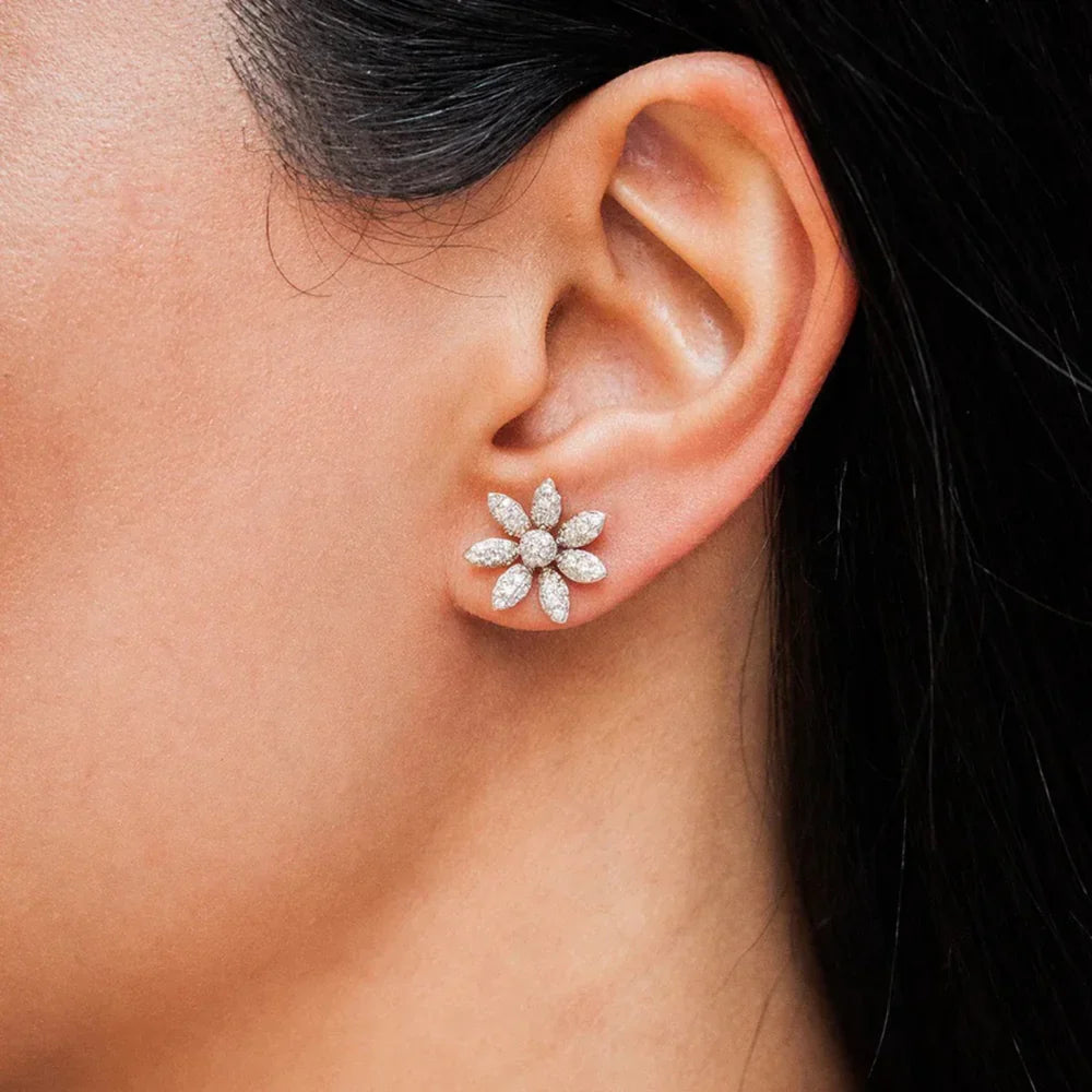 A close-up of a persons ear wearing a sparkling flower-shaped stud earring with multiple marquise-cut stones forming petals. The persons hair is dark and pulled back.